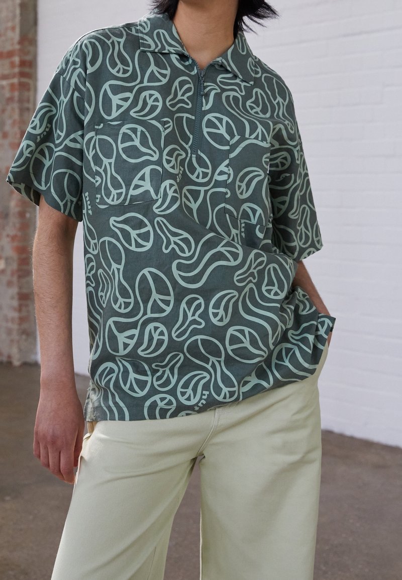 Camicia a maniche corte in tessuto verde scuro con motivi astratti di foglie verde chiaro, caratterizzata da un colletto e una tasca sul petto.