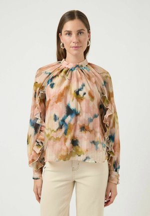 Vrouw met steil bruin haar die hoepeloorbellen draagt en een pastel meerkleurige blouse met ruches en lange mouwen en lichtbeige broek.