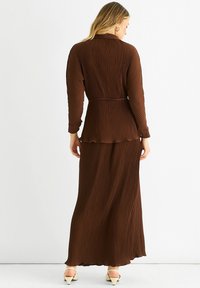 Brown pleated dress with long sleeves, belted waist, and flowing hem. Texture is ribbed; fabric has a slight sheen. Shoes are light-colored.
