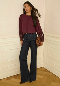 Vínová blúzka s čipkovými detailmi a nafúknutými rukávmi, kombinovaná s tmavými širokými džínsami. Modelka drží hnedé listové kabelku. Neutrálne pozadie.