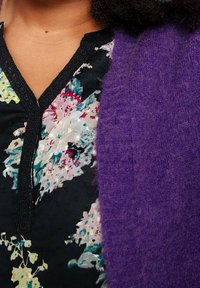 Blusa con motivo floreale su sfondo nero e accenti colorati, indossata sotto un cardigan viola morbido e lavorato a trama.