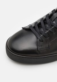 Černé kožené tenisky s hladkým, lesklým svrškem, plochými černými tkaničkami a texturovanou černou gumovou podrážkou; minimalistický design s funkčními detaily v šití.