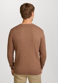 M�ężczyzna odwrócony plecami, ubrany w brązowy sweter w warkocze i beżowe spodnie, na jednolitym jasnoszarym tle.