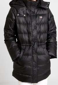 Abrigo acolchado negro con capucha, que presenta un acabado brillante, acolchado horizontal, botones a presión y un pequeño parche de marca en el pecho.