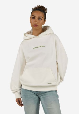 Kvinde med krøllet hår sat op i en knold, iført en hvid hættetrøje med teksten "Matcha on my mind" og lyseblå jeans, poserer foran en hvid baggrund.