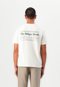 T-shirt en coton couleur crème avec un imprimé graphique vert au dos, ressemblant à un titre de journal, avec une coupe décontractée.