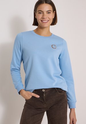 Femme souriante aux cheveux courts et bruns porte un sweat-shirt bleu clair et un pantalon marron foncé, debout avec une main dans la poche.