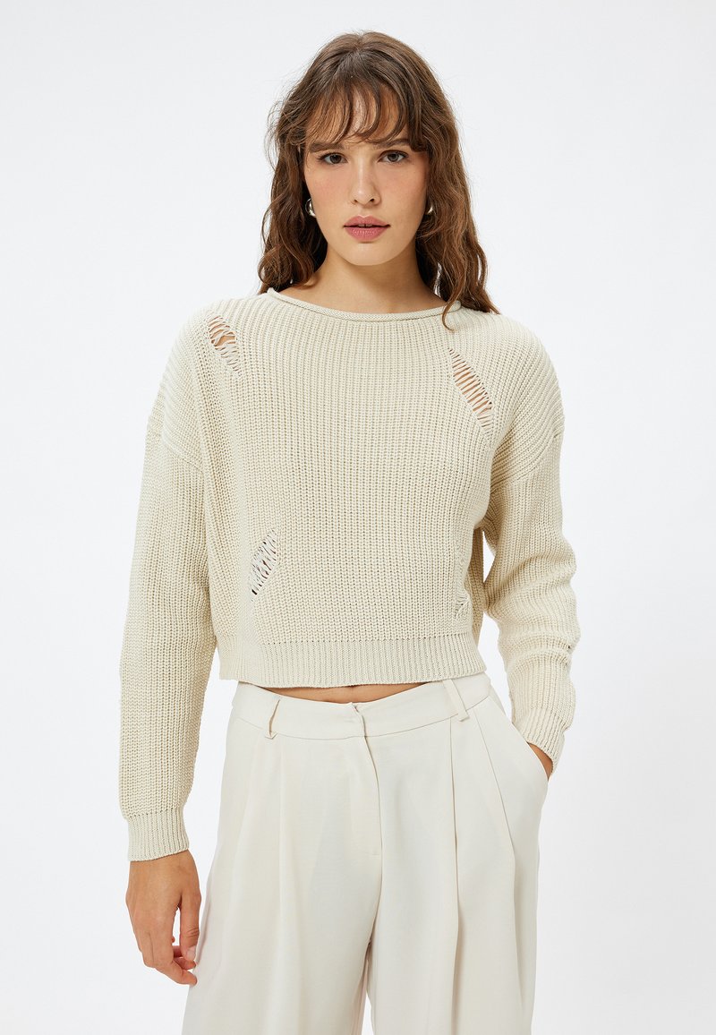 Gehaakte crèmekleurige trui met een cropped ontwerp, voorzien van versleten details en lange mouwen, gecombineerd met hoog-waist crèmekleurige broeken.