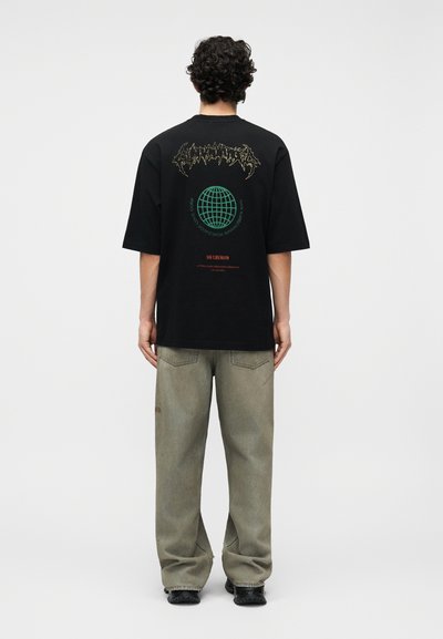 Άνδρας με σγουρά μαλλιά, ντυμένος με μαύρο γραφικό T-shirt με σχέδιο γης και κείμενο, συνδυασμένο με χαλαρά γκρι τζιν και μαύρα παπούτσια, στέκεται με την πλάτη προς τα εμπρός.