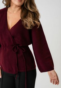 Femeie purtând un pulover tricotat de culoare burgundă adâncă, cu mâneci lungi legat la talie, asortat cu pantaloni negri, pe un fundal simplu.