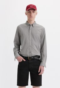 Camisa de botón gris claro, pantalones cortos de mezclilla negra, cinturón trenzado negro y gorra roja con el logo "LEVI'S". Atuendo limpio y casual en un modelo delgado.