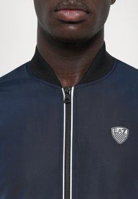 Casaco bombardeiro azul-marinho com um colarinho canelado preto, fecho à frente e riscas brancas de destaque. Apresenta um emblema no lado esquerdo.