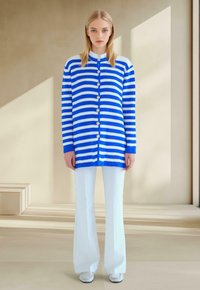 Cardigan rayé bleu et blanc avec des boutons, associé à un pantalon évasé blanc. Texture lisse et coupe décontractée. Détail de col simple.