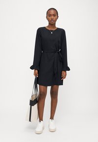 Μαύρο φόρεμα μακρύ μανίκι με φτερωτές μανσέτες και ζώνη στη μέση, κατασκευασμένο από ομαλό ύφασμα, φορεμένο με λευκά αθλητικά παπούτσια και μπεζ τσάντα χειρός.