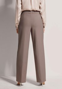 Lichtbruine wijde broek met een gladde textuur en elastische tailleband; gedragen met een lichtbeige zijdeachtige blouse, hakken zichtbaar eronder.