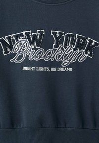 Tessuto blu scuro con ricamo in rilievo bianco e grigio con il testo "NEW YORK Brooklyn" e un testo più piccolo "LUCIDITÀ VIVIDE, GRANDI SOGNI."
