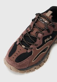 Zapatilla deportiva marrón y negra con materiales de ante y malla, cordones texturizados y una suela gruesa con acentos moteados. Logo de Fila visible.
