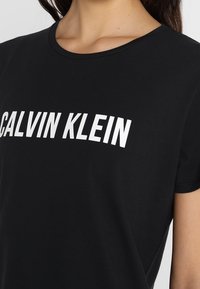 Camiseta negra de cuello redondo con el logo blanco "CALVIN KLEIN" en el pecho, llevada por una persona con cabello largo y oscuro.