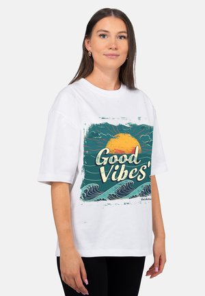 Young woman with long brown hair wearing white "Good Vibes" t-shirt featuring ocean waves and a setting sun graphic.