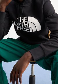 Persona seduta su uno sgabello, con una felpa nera con il logo "The North Face" e pantaloni verdi, mano appoggiata vicino al ginocchio.