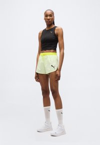 Γυναίκα που στέκεται φορώντας μαύρο αμάνικο τοπ και κίτρινα σορτς, με λευκές κάλτσες Puma και παπούτσια, σε απλό λευκό φόντο.