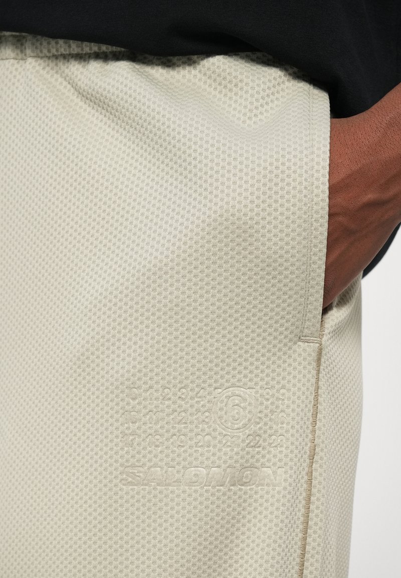 Shorts de sport beige clair en tissu texturé et respirant. Ils présentent un logo en relief et des détails numériques près de la poche.
