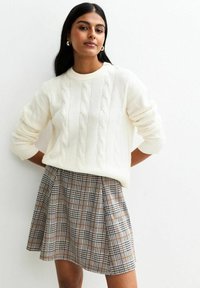 Frau in einem weißen Strickpullover und einem hochtaillierten grauen Karorock, die mit hinter dem Rücken verschränkten Händen vor einer schlichten weißen Wand steht.