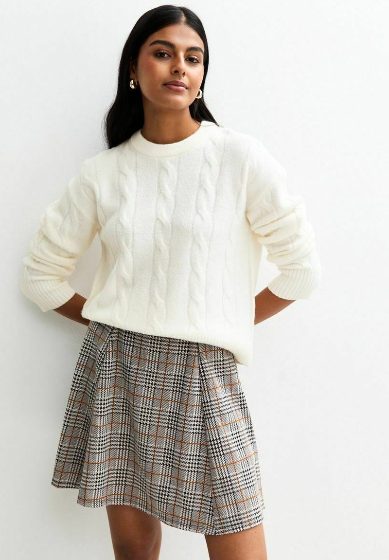 Frau in einem weißen Strickpullover und einem hochtaillierten grauen Karorock, die mit hinter dem Rücken verschränkten Händen vor einer schlichten weißen Wand steht.