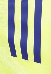 Helles gelbes Gewebe mit einem strukturierten Muster zeigt drei dunkelblaue vertikale Streifen. Das Material wirkt leicht und atmungsaktiv.