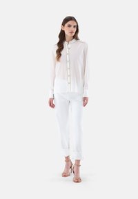 White long-sleeve blouse with a mandarin collar, featuring gold-toned button accents and a pocket, paired with white cuffed trousers and beige heeled sandals.