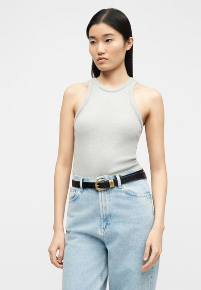 Νέα γυναίκα με μακριά σκούρα μαλλιά που φοράει αμάνικο ριμπ γκρι ανοιχτό τοπ, ψηλόμεσο ανοιχτό μπλε τζιν και μαύρη ζώνη με χρυσή αγκράφα.