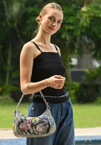 Graue Handtasche mit floralen Mustern, Kettenriemen, geschwungener Form und weicher Textur. Model trägt ein schwarzes Oberteil und Jeans, im Freien.