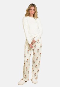 Langärmliges cremefarbenes Oberteil kombiniert mit cremefarbenen Hosen, die mit einem floralen Muster in Orange und Blau verziert sind, sowie weichen, plüschigen Hausschuhen.