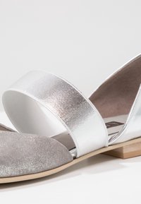 Gros plan d'une chaussure plate argentée avec un bout scintillant, une large bride métallique sur le dessus et un petit talon en bois.