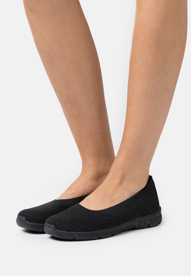 Jambes portant des chaussures de type slip-on en tricot noir avec des semelles en caoutchouc flexibles, debout sur une surface blanche.