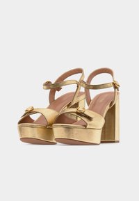 Χρυσά μεταλλικά πέδιλα με χοντρά τ ακούνια, λουράκια αστραγάλου, ανοιχτά δάχτυλα και κλείσιμο με πόρπη, σε απλό φόντο.