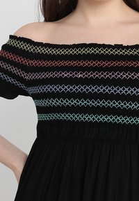 Mustassa off-shoulder-mekossa on monivärisiä brodeerattuja raitoja tyylikkään yläosan ympärillä. Materiaalissa on strukturoidut smokkityyliset yksityiskohdat ja vyötärön alapuolella hulmuava kangas.