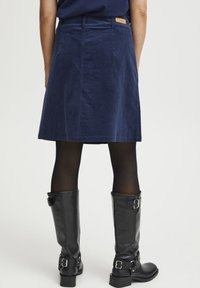 Gonna A-line in corda navy con vita e etichetta, abbinata a stivali neri in pelle al ginocchio con fibbie e finitura testurizzata.