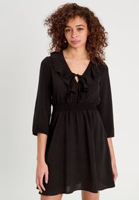 Robe noire dotée d'un décolleté en V avec des volants, d'un nœud au niveau du cou, de manches trois-quarts et d'une taille élastique, en tissu léger.