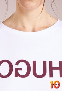 Camiseta blanca con texto en marcón audaz "HUGO" en una fuente moderna, con un cuello redondo y una textura de tela suave. "10 AÑOS" en acentos coloridos.