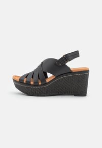 Sandales noires à talon compensé avec un design à lanières, tige en cuir, semelle en bois et semelle noire texturée. Sangle réglable à l'arrière.