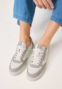 Adidașii au un superior din piele albă cu accente din suede gri, vârf rotund și închidere cu șireturi; design minimalist cu suprafață texturată.