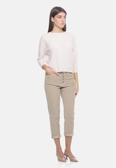 Donna in piedi con una mano in tasca, che indossa un maglione rosa chiaro, pantaloni cropped beige con bottoni e tacchi beige su sfondo bianco.