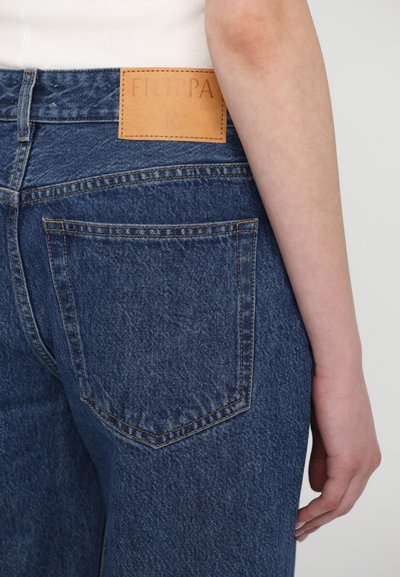 Vaqueros de mezclilla azul con un parche de cuero etiquetado "FILIPPA K" en la cintura, que cuentan con bolsillos traseros y detalles de costura visibles.