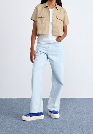 Persona con una camisa beige corta abotonada, top blanco, vaqueros de pierna ancha azul claro y zapatillas plataforma azules de pie sobre un suelo gris.