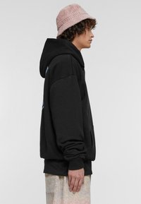 Sudadera negra con capucha y mangas largas, bolsillo frontal y puños de canalé, combinada con un sombrero tipo cubo texturizado en color rosa.