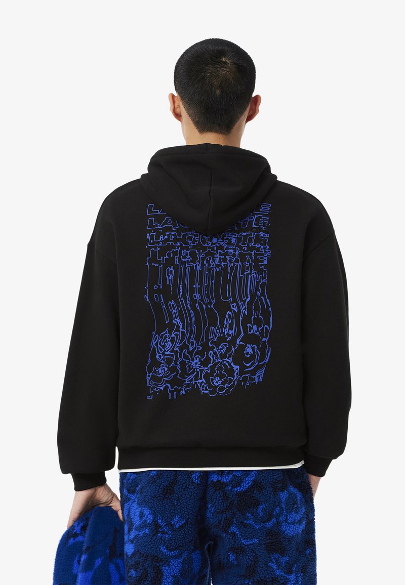 Felpa nera con un design grafico blu sulla schiena, caratterizzato da motivi astratti. Il modello indossa pantaloni blu con texture e tiene un oggetto coordinato.