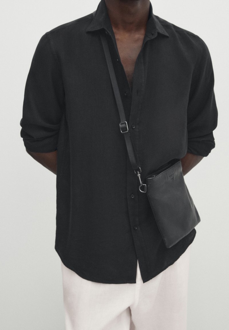 Homme portant une chemise noire boutonnée et un pantalon blanc avec un petit sac noir en bandoulière suspendu sur la poitrine.