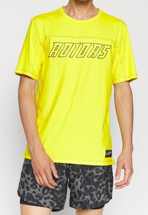 Mand iført en lysegul Adidas T-shirt og sorte-grå mønstrede træningsshorts står med armene afslappede.