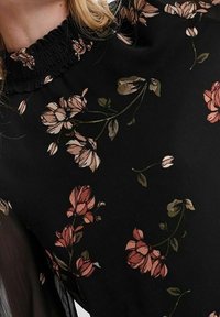 Blusa nera con motivo floreale e scollo arricciato. Presenta fiori beige e rosa su un tessuto texturizzato. Maniche lunghe con un design fluido.